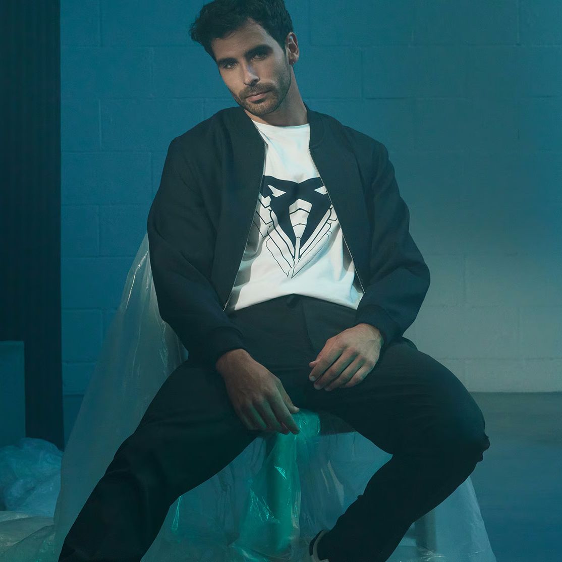 Oseba v črnem suknjiču in beli majici kolekcije CUPRA COLLECTION. Na majici je velik logotip CUPRA. Oseba sedi v temnem prostoru z modrimi toni v ozadju..