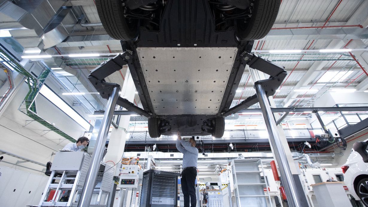 Mehanik dela na podvozju dvignjenega avtomobila v svetli delavnici, obkrožen z orodjem in opremo. Zgornji del delavnice je viden z večjimi okni in fluorescentno osvetlitvijo. 