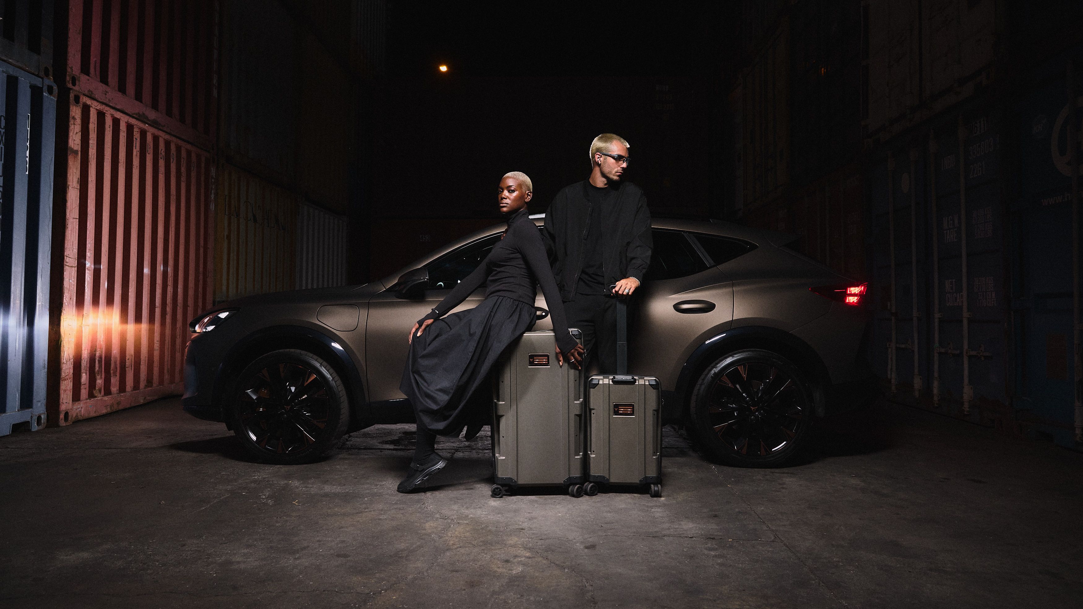 Dve osebi stojita poleg bronastega avtomobila v slabo osvetljenem prostoru s kovčki pred seboj.