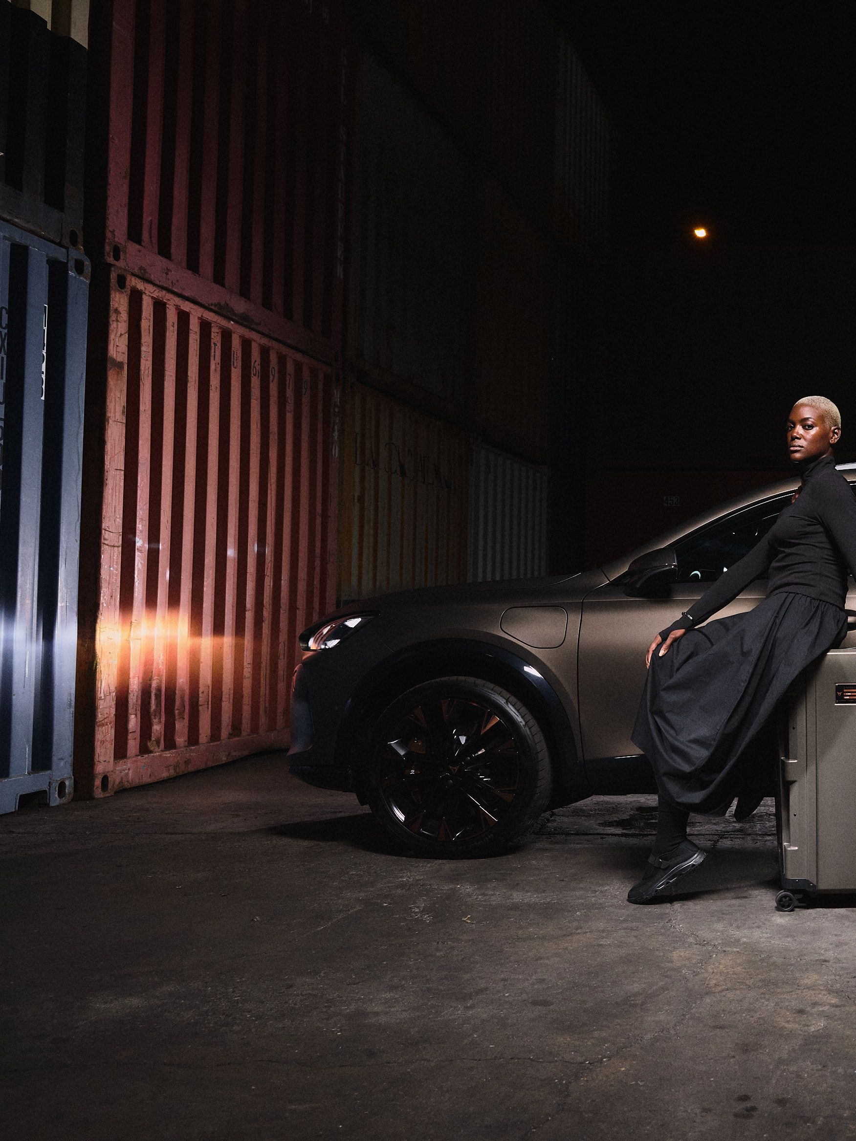 Dve osebi stojita poleg bronastega avtomobila v slabo osvetljenem prostoru s kovčki pred seboj.