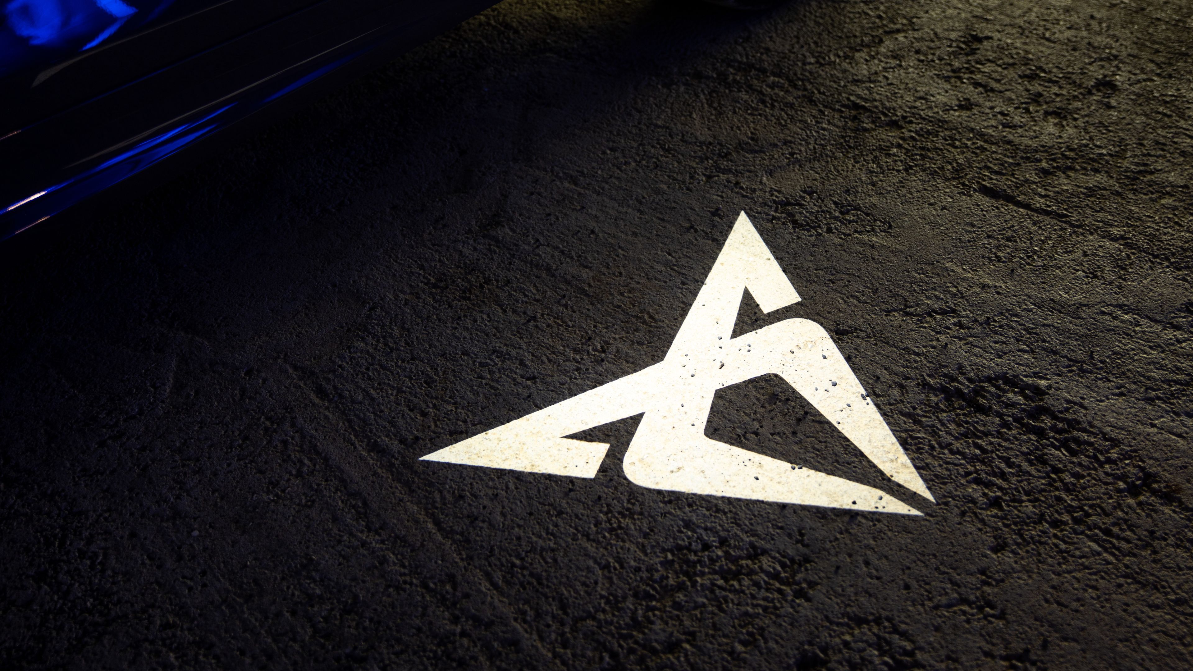 Osvetljen logotip znamke CUPRA na asfaltni podlagi, ki ga projicira LED lučka iz avtomobila. Podlaga je groba in temna, kar poudarja svetlost projekcije.