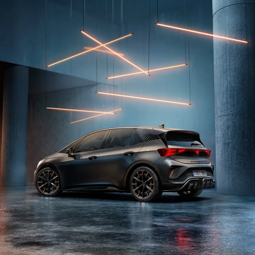 Temno siv CUPRA Born stoji v velikem industrijskem prostoru. Nad avtom visijo tanki oranžni svetlobni trakovi, ki ustvarjajo futuristično vzdušje. 