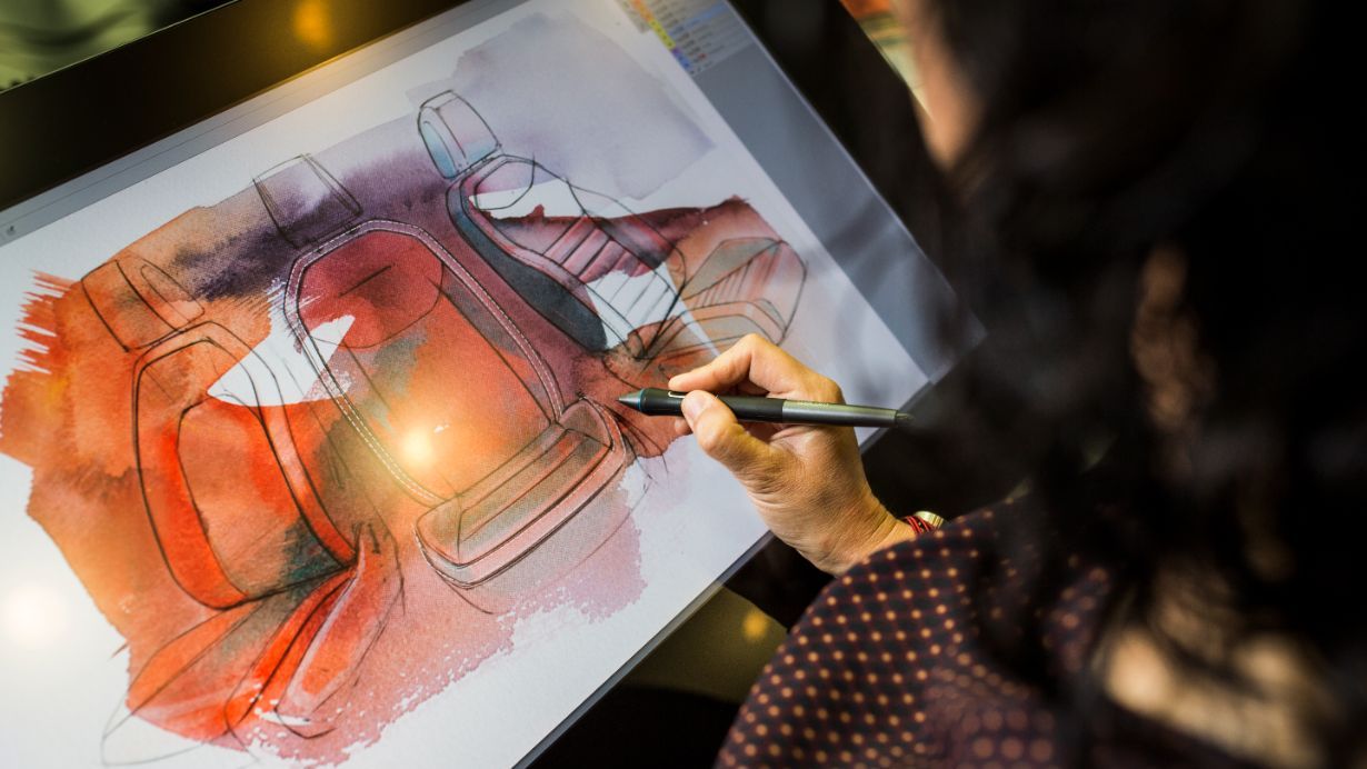 Oseba na risalni tablici ustvarja barvno skico avtomobilskih sedežev. Skica vključuje rdeče in oranžne odtenke. Oseba drži pisalo in riše na digitalnem zaslonu.