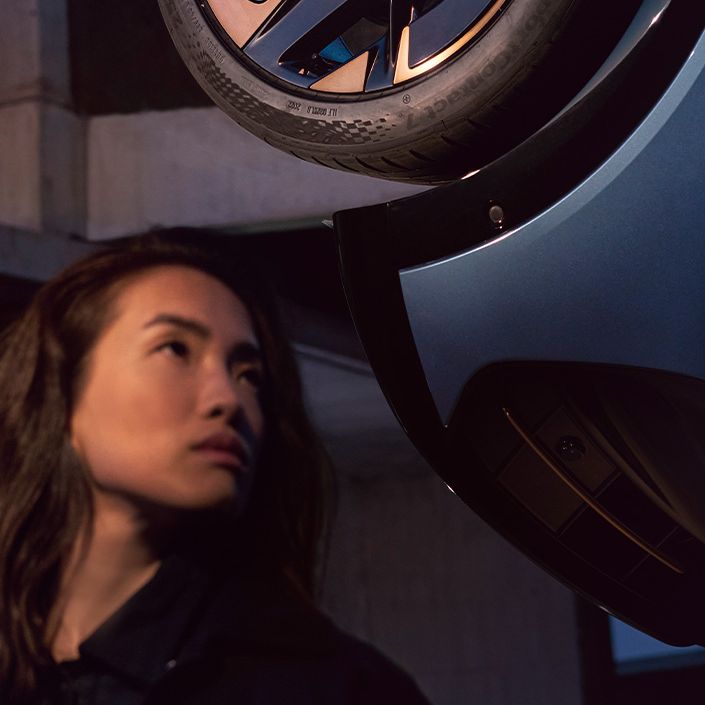 V dolgem navpičnem kadru je prikazana pnevmatika Cupre Tavascan, ki je dvignjen. Pod avtomobilom stoji ženska z zamegljenim obrazom.