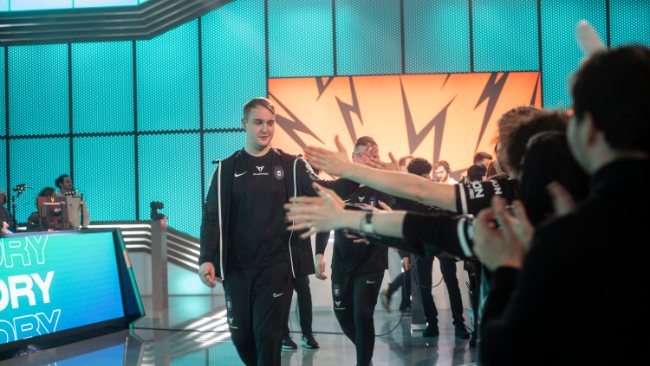 Skupina ljudi v športnih oblačilih hodi po odru, obkroženem z modrimi lučmi in velikim oranžnim zaslonom. Občinstvo na strani odra iztegne roke proti njim. 