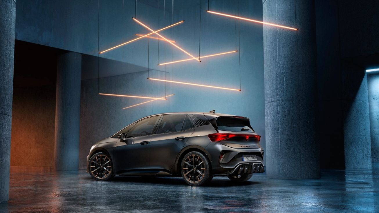Temno siv CUPRA Born stoji v velikem industrijskem prostoru. Nad avtom visijo tanki oranžni svetlobni trakovi, ki ustvarjajo futuristično vzdušje. 