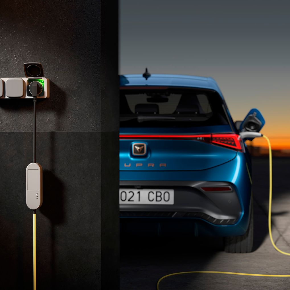 Električni avto CUPRA Born je priključen na polnilnico, ki je pritrjena na temen zid. Polnilnica ima več vtičnic, ena izmed njih je povezana s kablom, ki polni avto. V ozadju je sončni zahod.