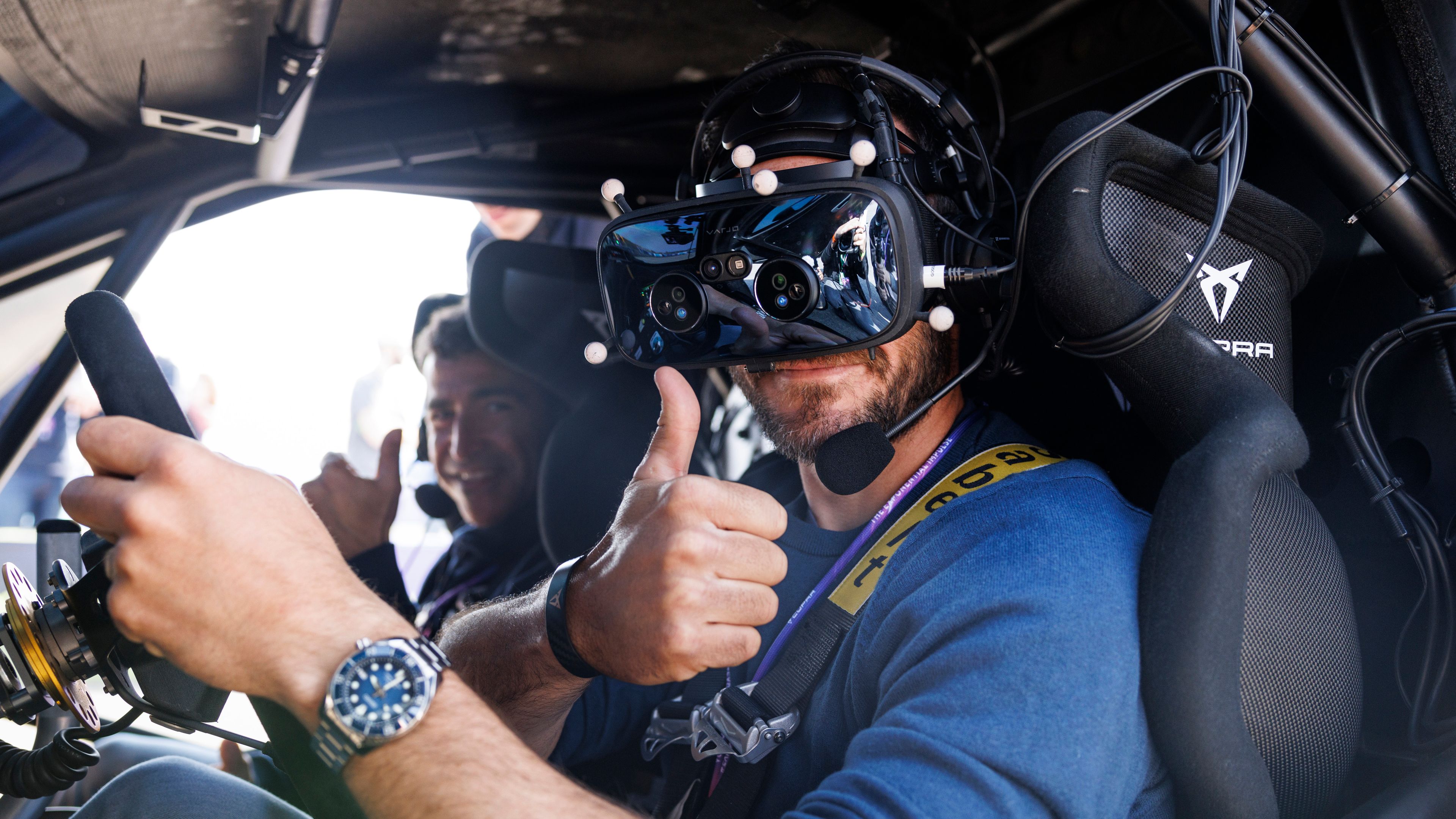 Dva moška sedita v dirkalniku znamke CUPRA. Moški, ki sedi na voznikovem sedežu, nosi VR-očala in kaže palec navzgor.