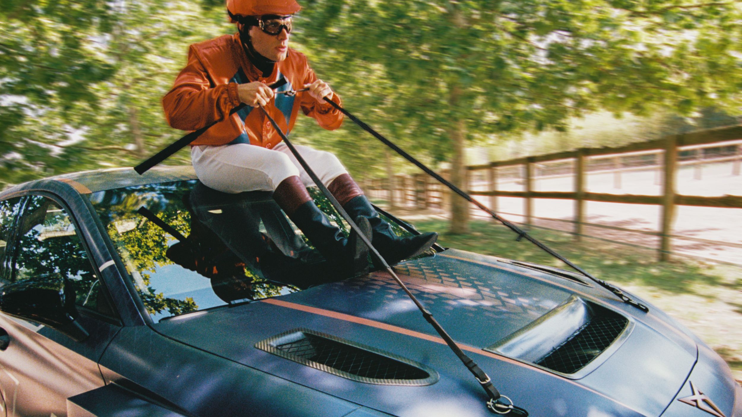 Oseba v jahalni opremi z oranžno čelado in očali sedi na pokrovu modrega športnega avtomobila ter drži vajeti, kot da jezdi avto, med vožnjo po drevoredni cesti.