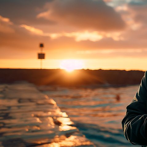 Moški z brado stoji ob morju ob sončnem zahodu in gleda v mobilni telefon, kar simbolizira povezljivost in interakcijo posameznika s tehnologijo na prostem.