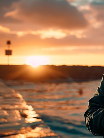 Moški z brado stoji ob morju ob sončnem zahodu in gleda v mobilni telefon, kar simbolizira povezljivost in interakcijo posameznika s tehnologijo na prostem.
