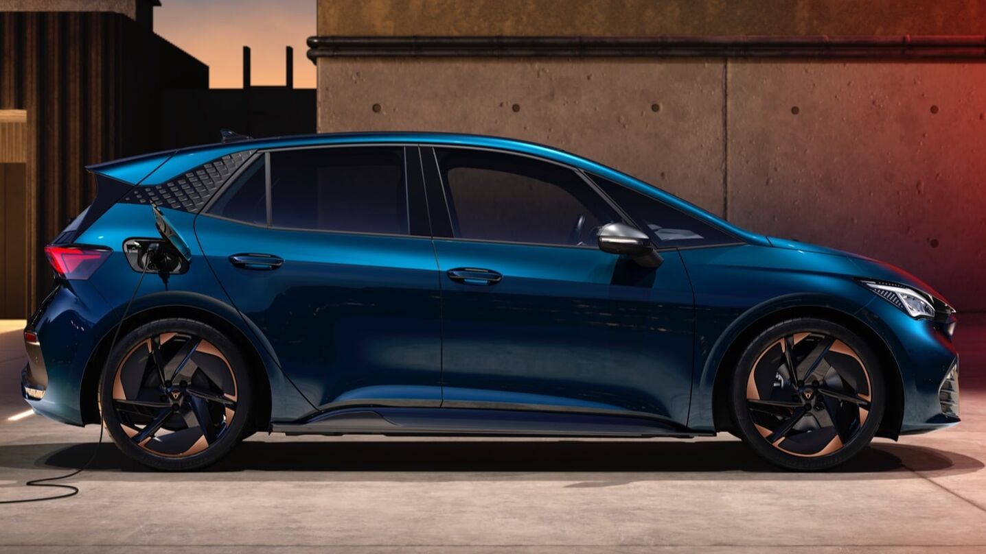 Električni avtomobil CUPRA Born modre barve se polni. Vozilo je parkirano ob steni z vkjučenim kablom za polnjenje. Ozadje je betonska stena in sončni zahod.