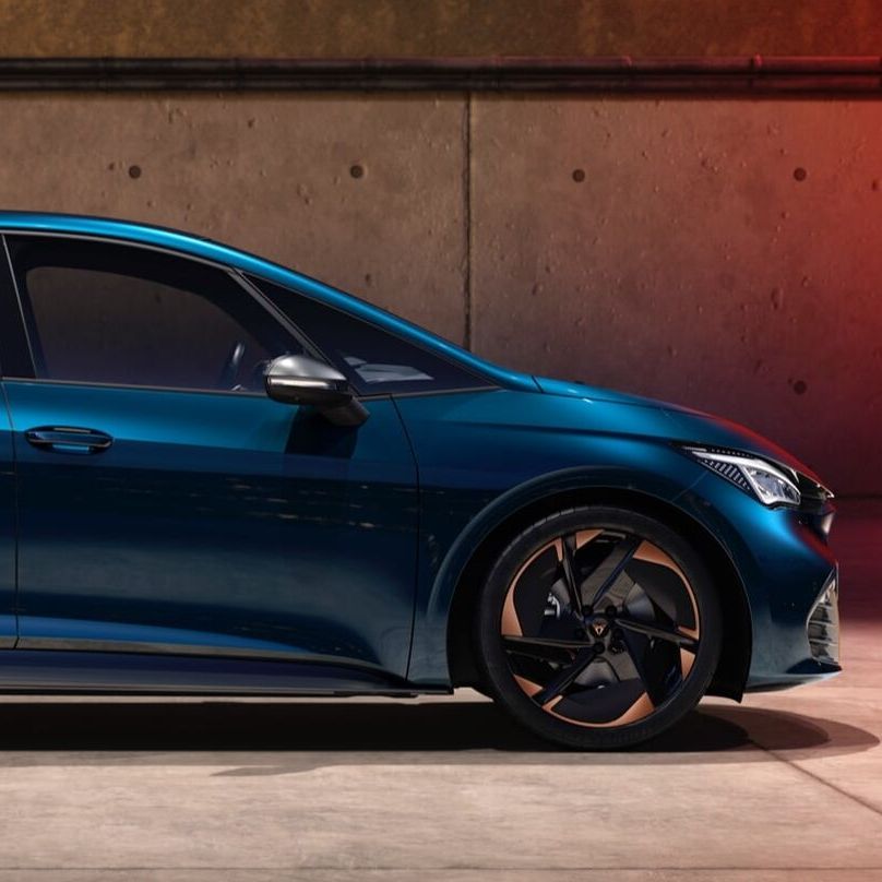 Električni avtomobil CUPRA Born modre barve se polni. Vozilo je parkirano ob steni z vkjučenim kablom za polnjenje. Ozadje je betonska stena in sončni zahod.