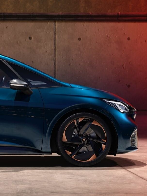 Električni avtomobil CUPRA Born modre barve se polni. Vozilo je parkirano ob steni z vkjučenim kablom za polnjenje. Ozadje je betonska stena in sončni zahod.