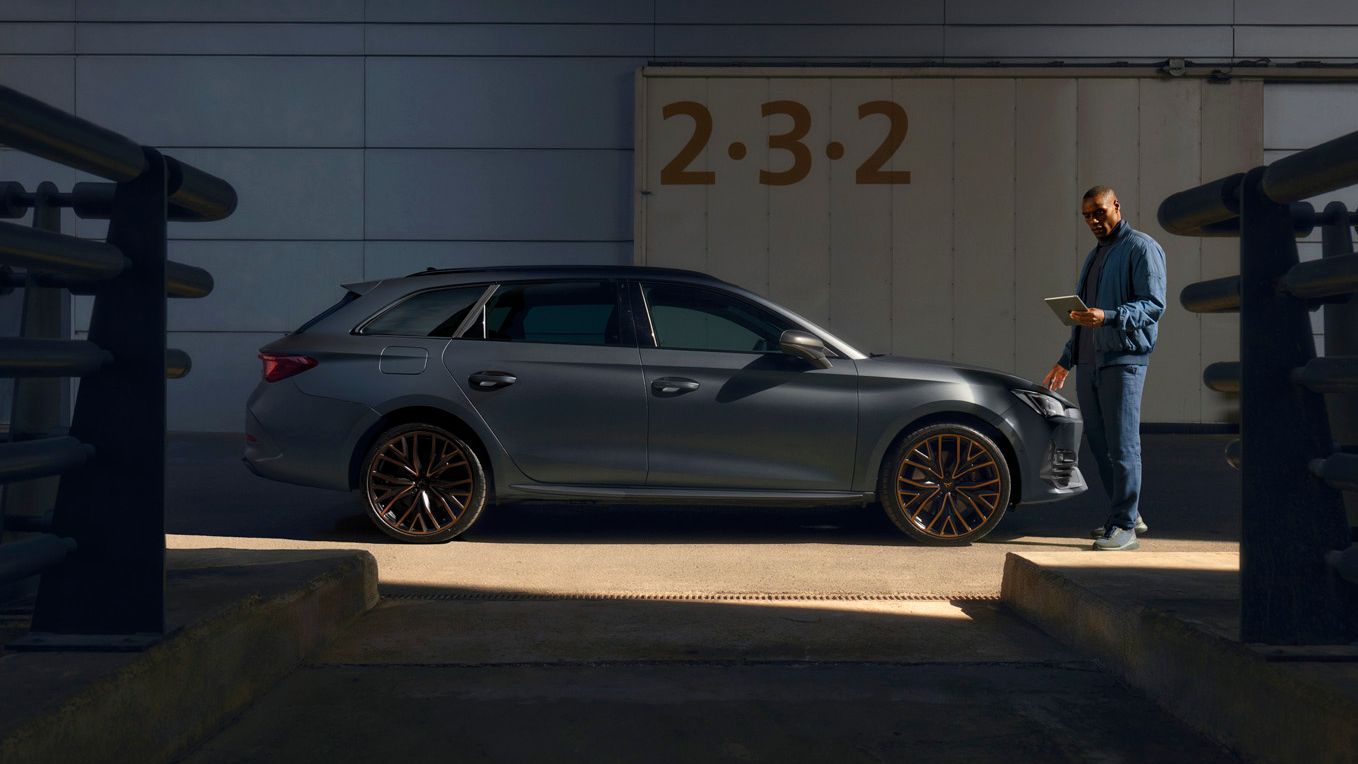 Moški stoji ob temnem karavanu CUPRA Leon SP, parkiranem pred modernim stavbnim vhodom.