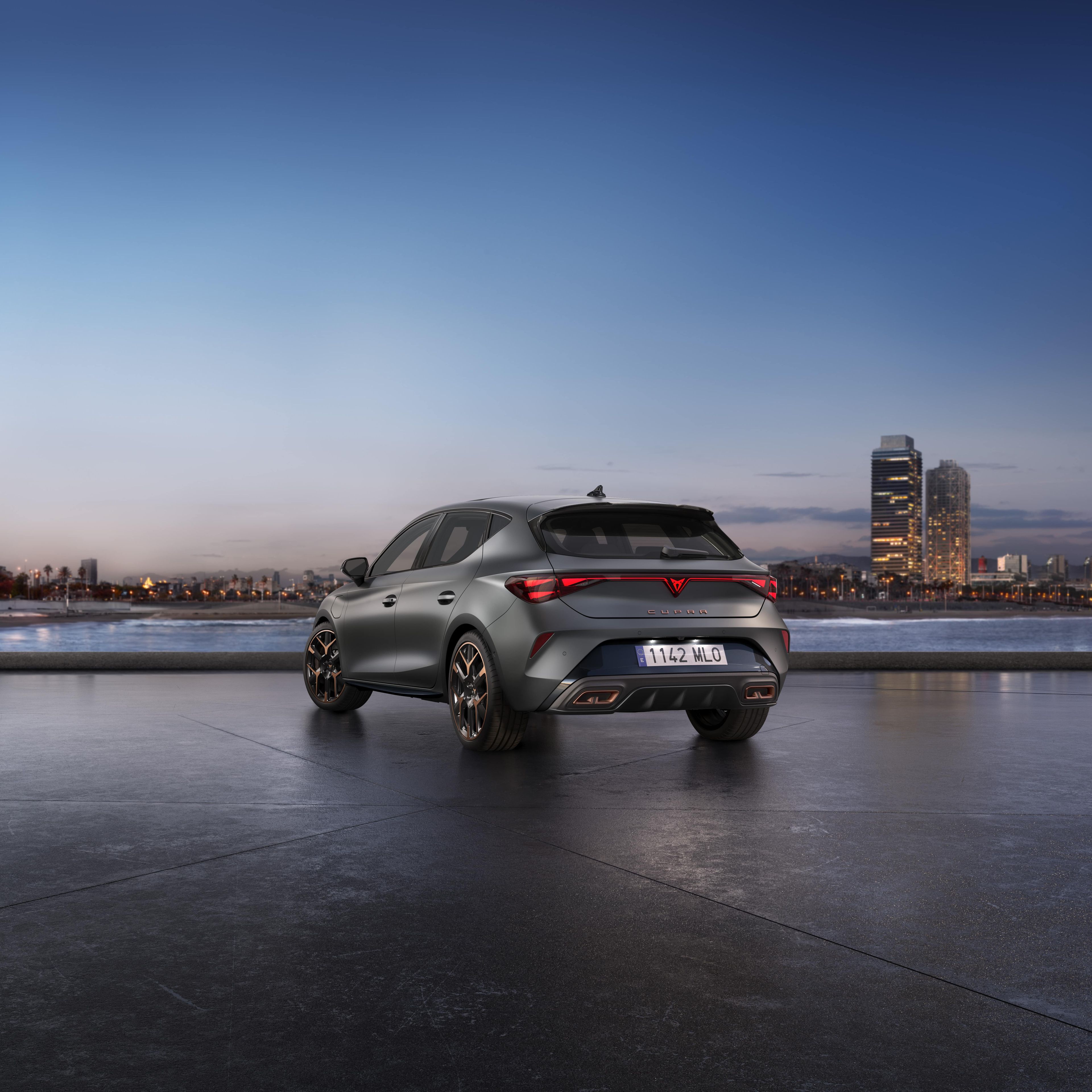 Cupra Leon VZ je parkiran na obali ob mestu, ob sončnem zahodu. V ozadju se vidi mesto z visokimi zgradbami. Nebo je jasno z odtenki modre in oranžne barve.