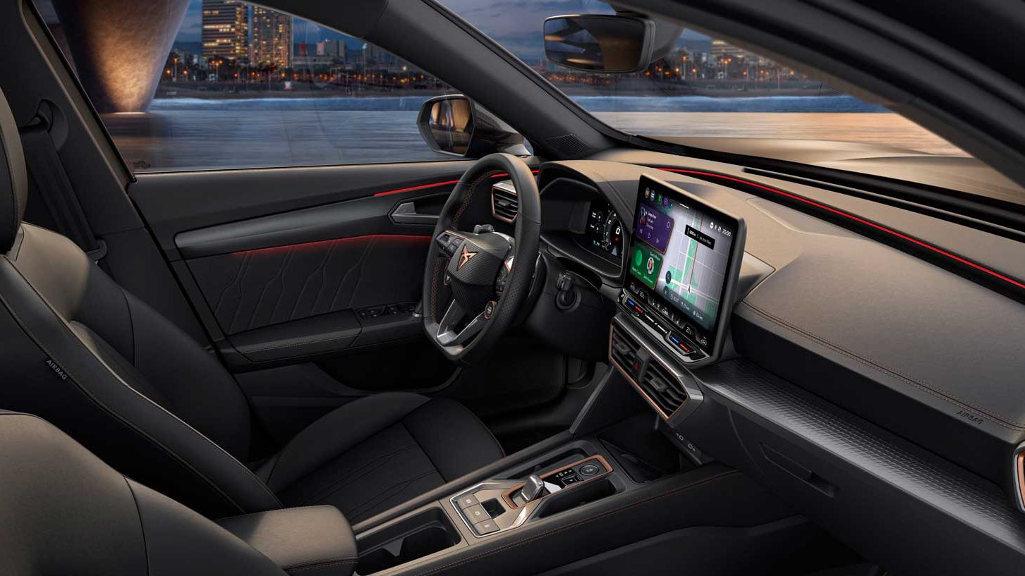 Notranjost sodobnega vozila CUPRA Formentor z velikim zaslonom infotainment sistema, volanom in upravljalnimi elementi. Vozilo ima elegantne črne sedeže, skozi okna pa se vidi osvetljeno mesto.