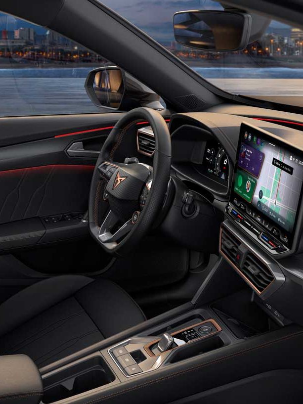 Notranjost sodobnega vozila CUPRA Formentor z velikim zaslonom infotainment sistema, volanom in upravljalnimi elementi. Vozilo ima elegantne črne sedeže, skozi okna pa se vidi osvetljeno mesto.