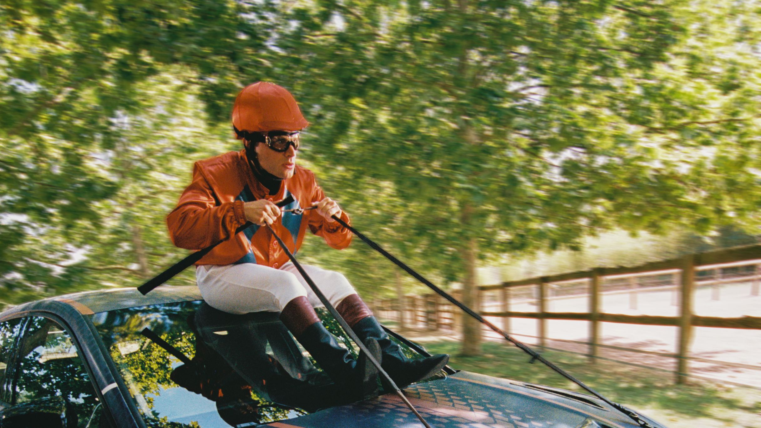 Oseba v jahalni opremi z oranžno čelado in očali sedi na pokrovu modrega športnega avtomobila ter drži vajeti, kot da jezdi avto, med vožnjo po drevoredni cesti.