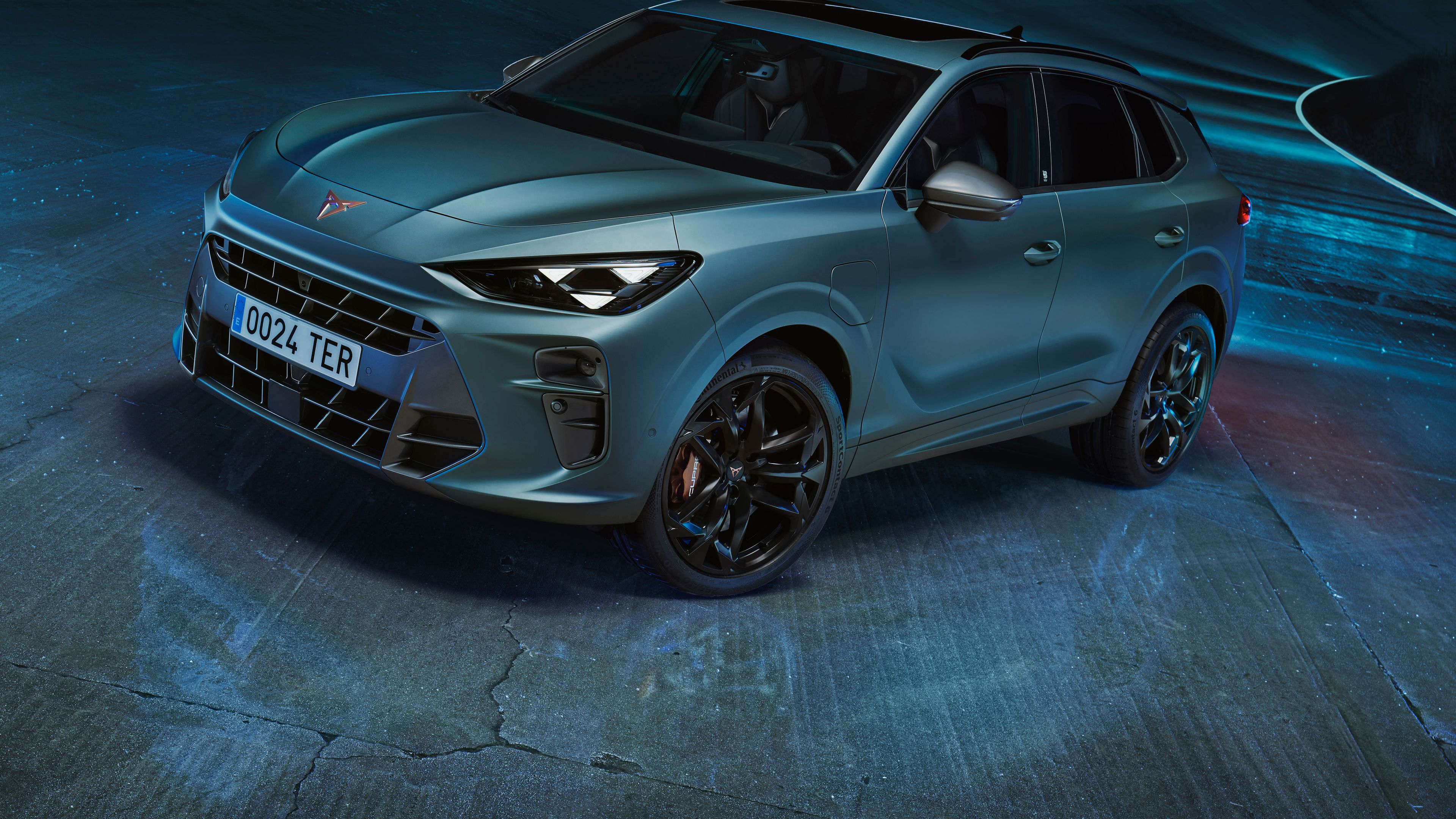 Kovinsko modri mat CUPRA Terramar z osvetljenimi žarometi stoji na modernem, osvetljenem cestnem ovinku. Parkiran je na mokrem betonu, ki poudarja njegov sodoben in dinamičen videz.