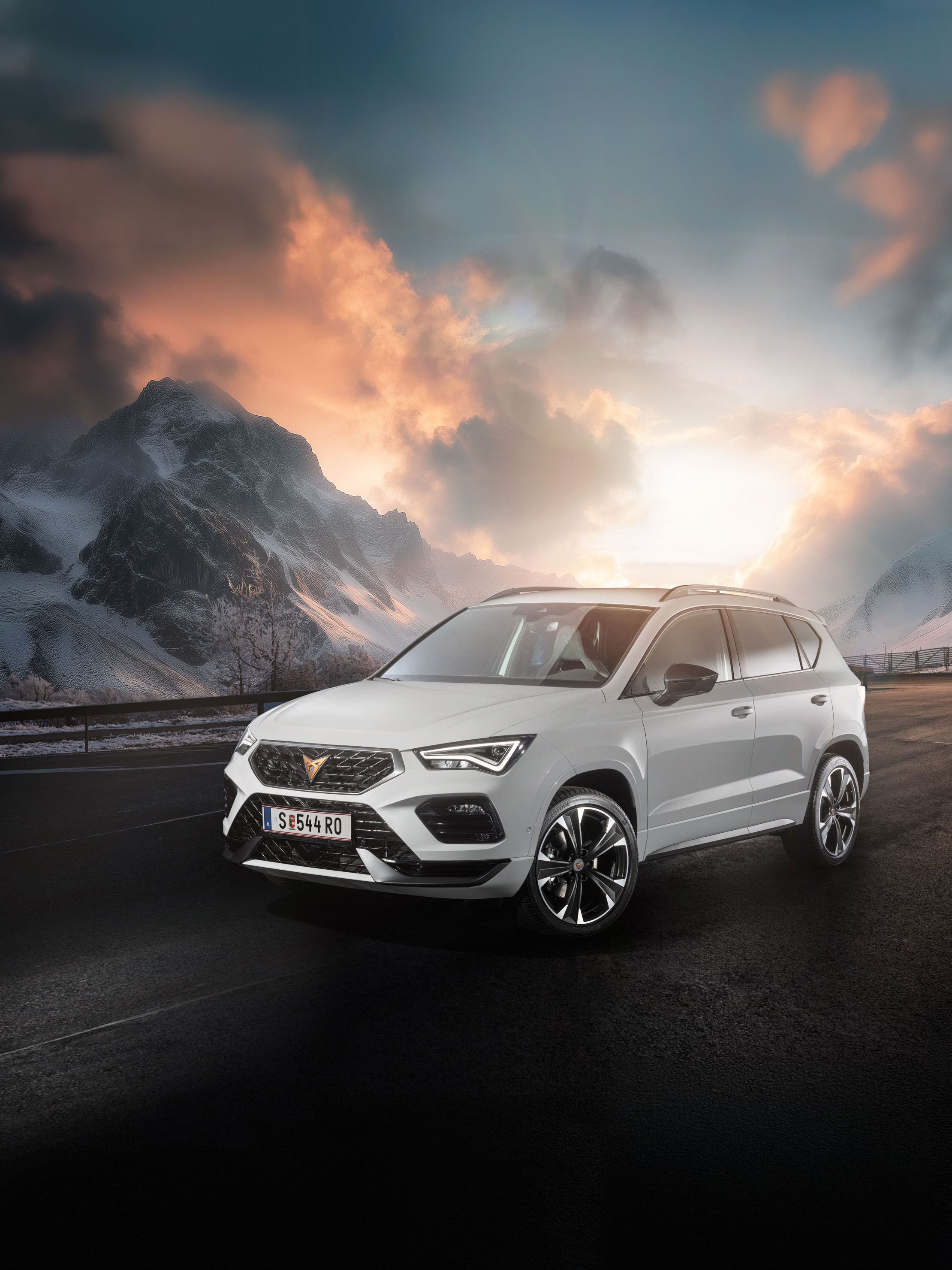 Ein weißer CUPRA Ateca fährt auf einer Bergstraße bei Sonnenaufgang. Die Straße ist nass und von Schnee gesäumt. Im Hintergrund sind schneebedeckte Berge und bewölkter Himmel zu sehen.