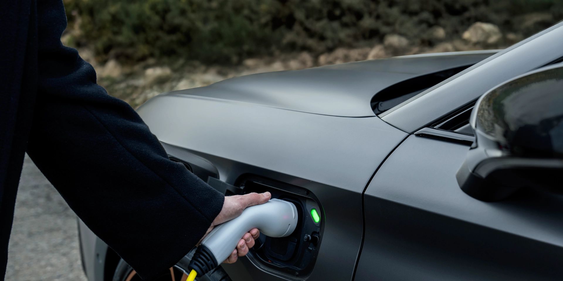 Oseba v temnem plašču priključuje polnilni kabel v priključek električnega avtomobila CUPRA Leon.