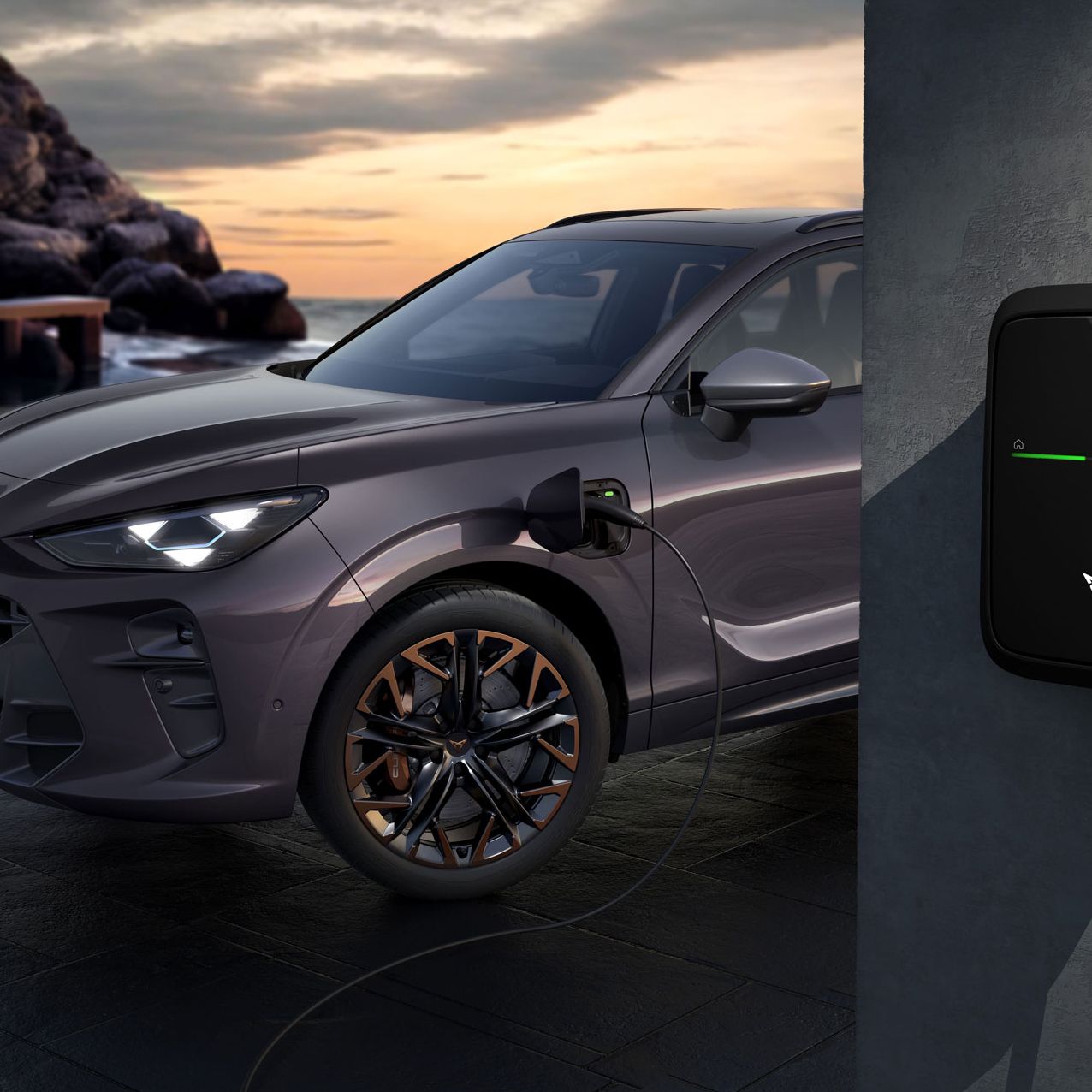 Električno vozilo CUPRA Terramar PHEV VZ, je priključeno na polnilno postajo na zunanjem dvorišču ob sončnem zahodu. Avto je vijolično-siv in stoji ob moderni polnilni enoti na steni. 