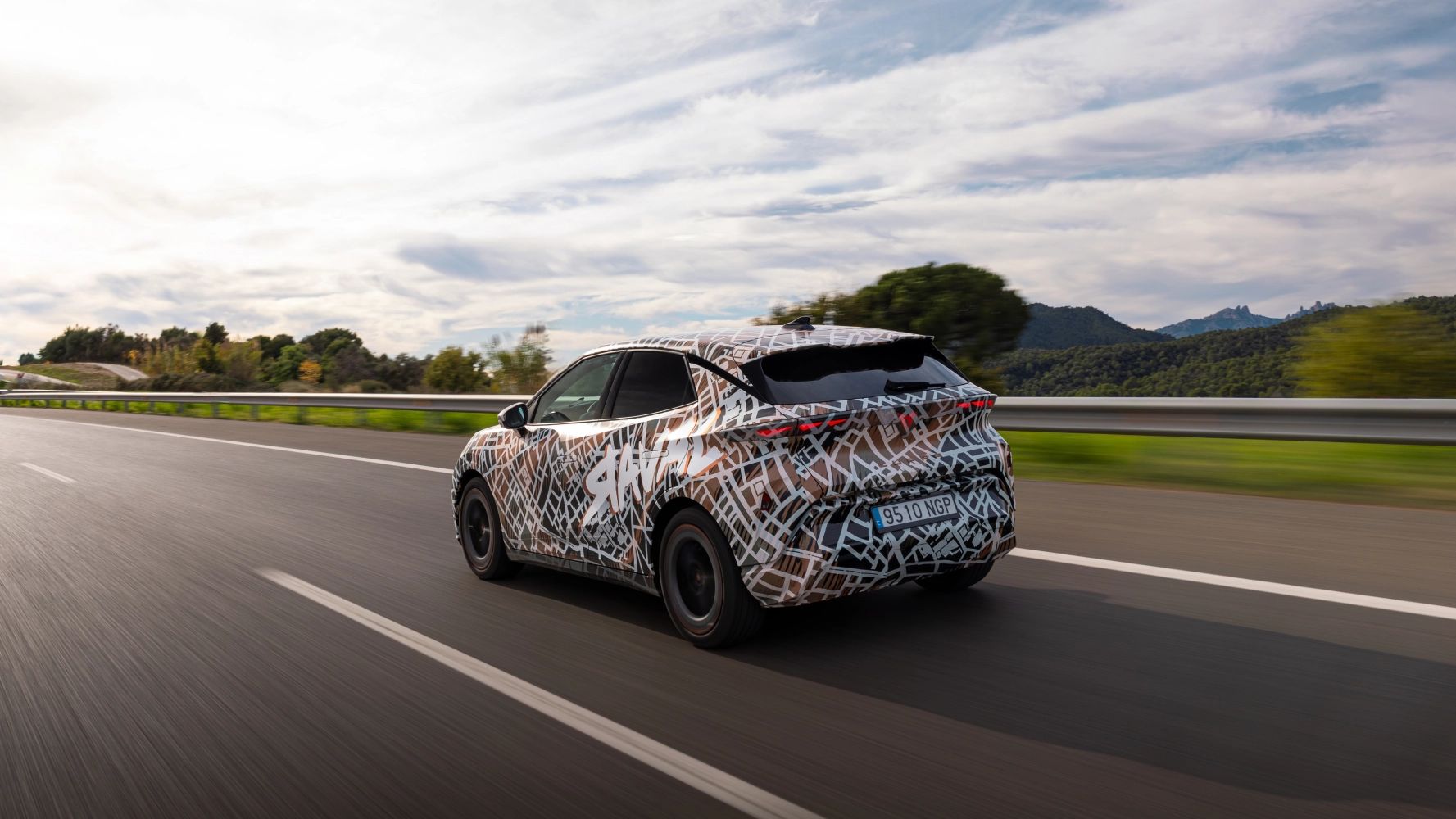Zamaskiran prototip avtomobila CUPRA Raval, se pelje po avtocesti v naravnem okolju s hribovjem v ozadju. Nebo je delno oblačno.