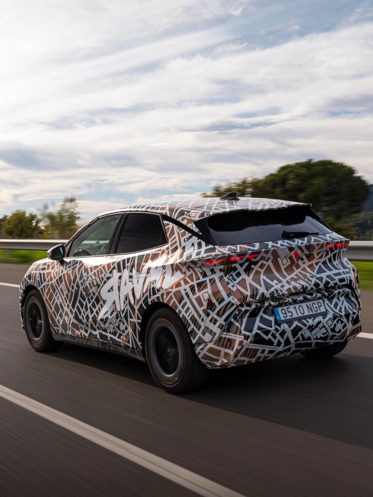 Zamaskiran prototip avtomobila CUPRA Raval, se pelje po avtocesti v naravnem okolju s hribovjem v ozadju. Nebo je delno oblačno.