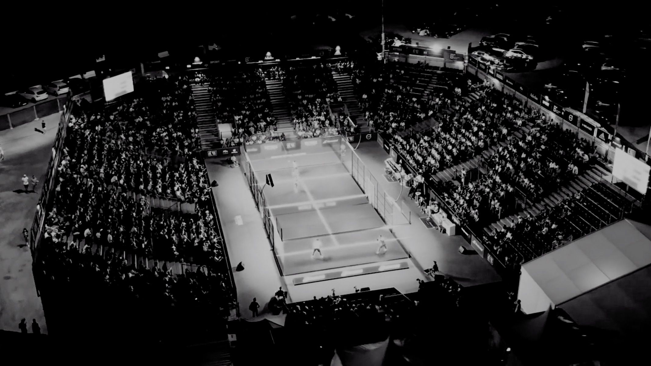 Črno-bel posnetek prikazuje turnir padel tenisa, s številnimi gledalci v tribunah ter osvetljeno igrišče.