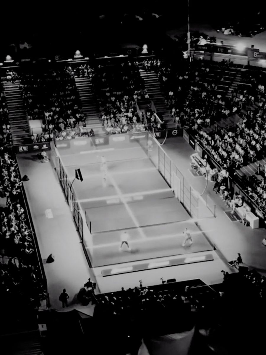 Črno-bel posnetek prikazuje turnir padel tenisa, s številnimi gledalci v tribunah ter osvetljeno igrišče.