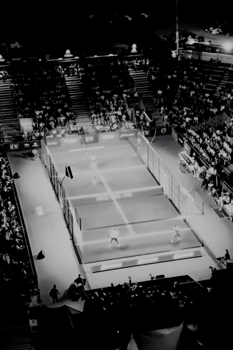 Črno-bel posnetek prikazuje turnir padel tenisa, s številnimi gledalci v tribunah ter osvetljeno igrišče.