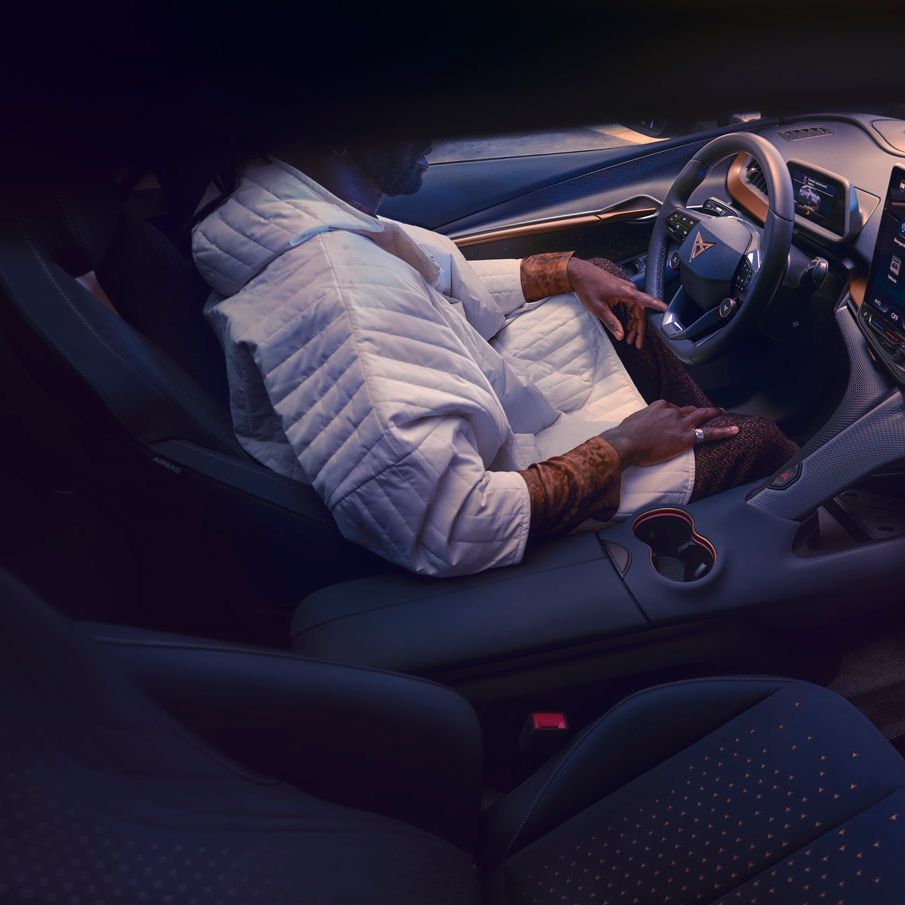 Notranjost avtomobila CUPRA Tavascan VZ, kjer oseba sedi na voznikovem sedežu in uporablja velik zaslon na dotik na armaturni plošči. Armaturna plošča ima minimalističen dizajn z digitalnimi prikazovalniki.