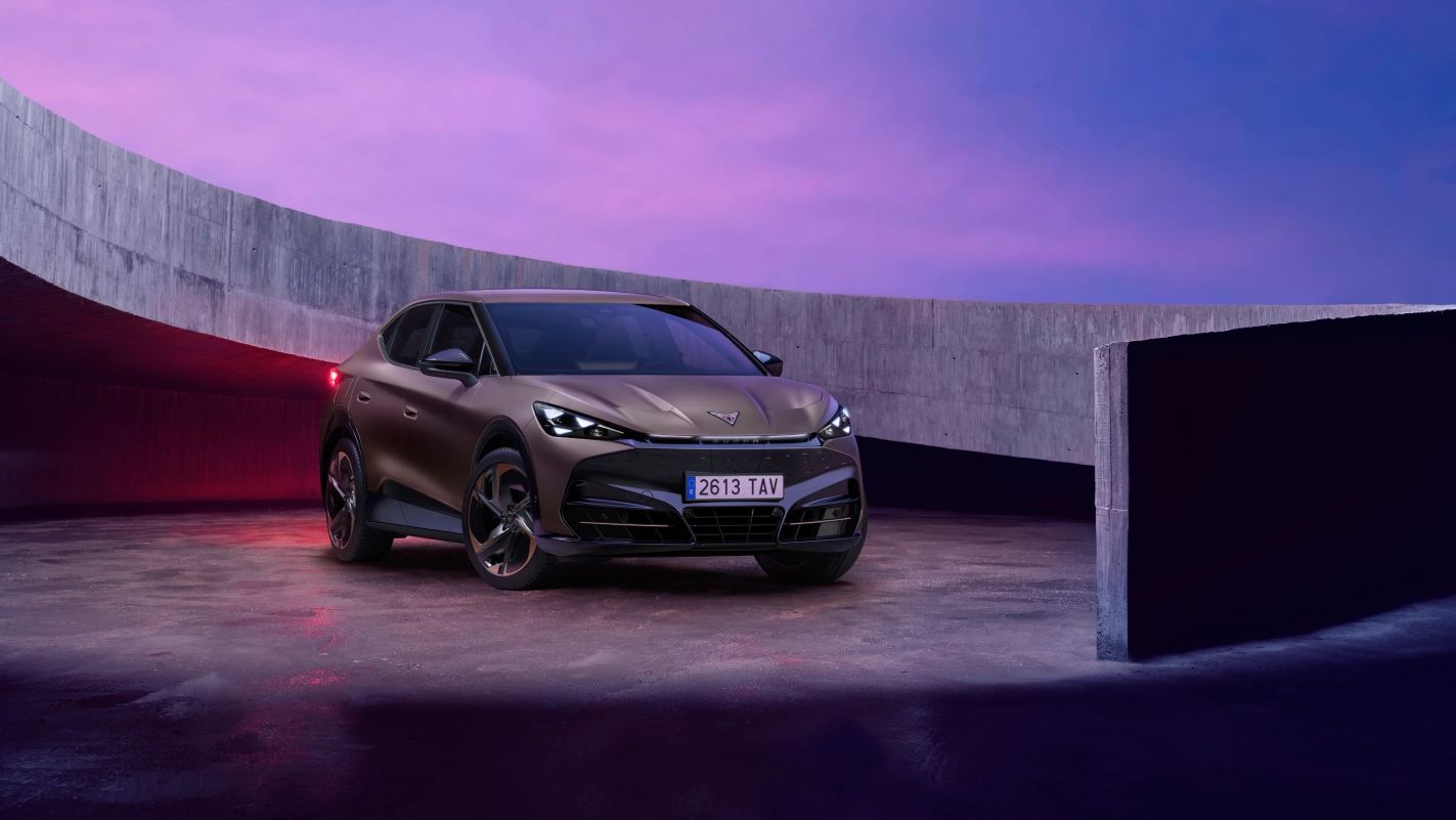 CUPRA Tavascan stoji na betonski rampi z elegantnim, ukrivljenim zidom. Ozadje je osvetljeno z dramatičnim vijoličnim in rožnatim nebom.