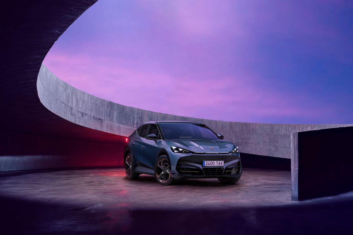 CUPRA Tavascan VZ je parkiran pod ovinkom betonske strukture na ploščadi ob sončnem zahodu. Avto je temno modre barve z modernim dizajnom.