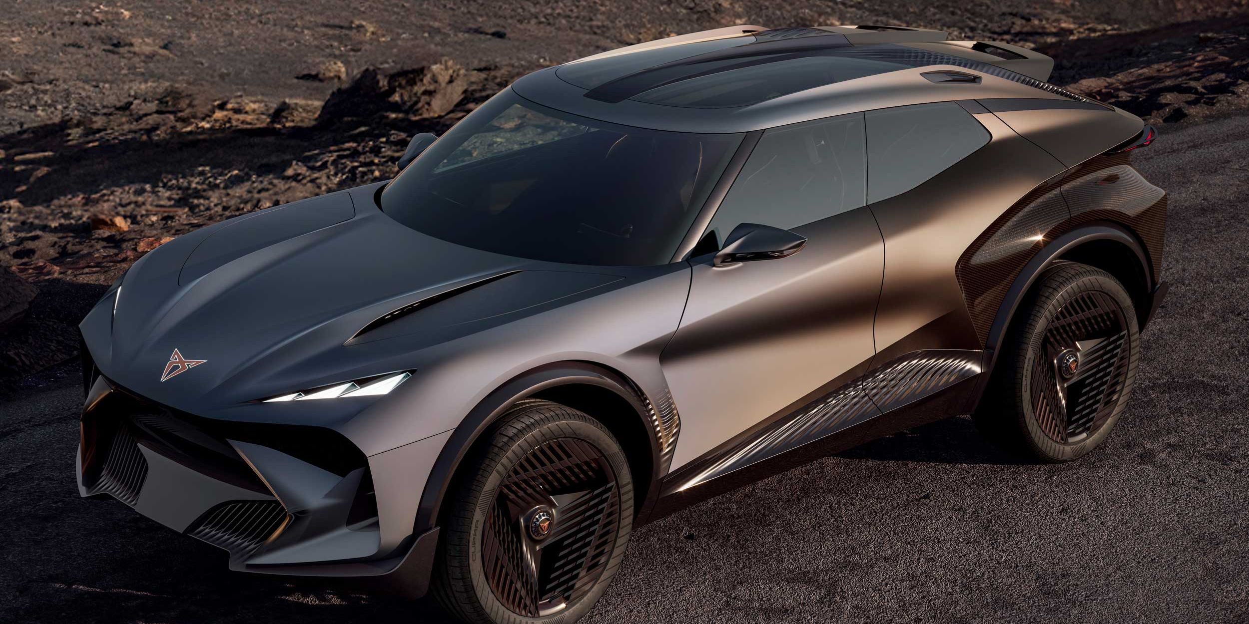 Futurističen športni SUV s kovinsko sivo karoserijo z ostrimi linijami in velikimi črnimi platišči vozi po prazni, asfaltirani cesti. V ozadju je surova, kamnita, skoraj vulkanska pokrajina brez vegetacije. Avto deluje kot konceptno vozilo z agresivnim dizajnom in poudarjenimi aerodinamičnimi elementi.
