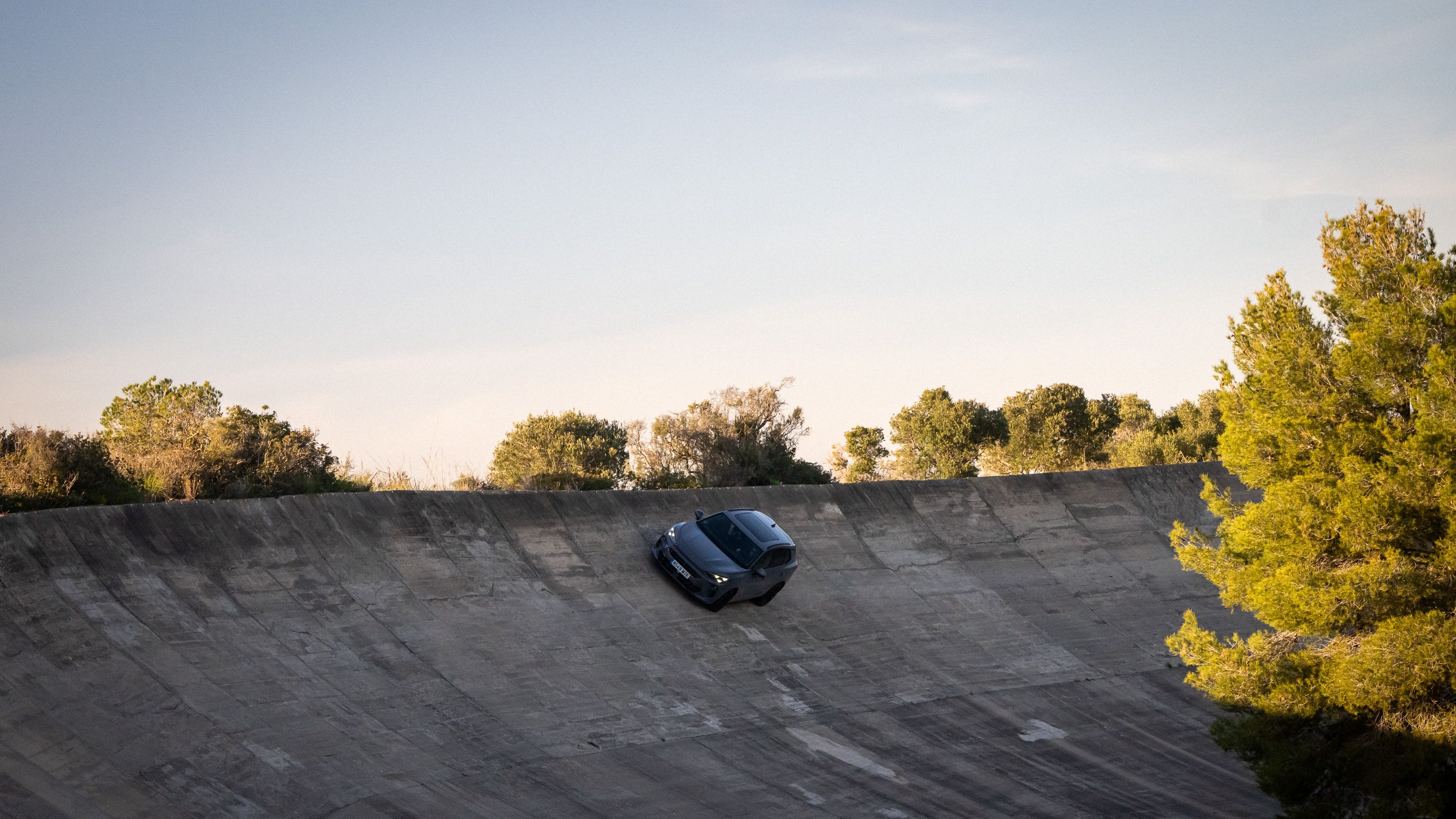 Športni avto CUPRA Terramar vozi po strmem, ukrivljenem betonskem površju, obsijan z zadnjimi sončnimi žarki. V ozadju so drevesa in grmičevje.