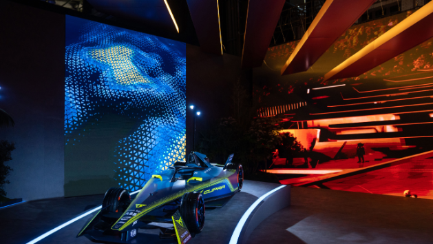 Dirkalnik z modernim dizajnom in svetlobnimi efekti je razstavljen v osvetljenem prostoru. Ozadje je dinamično s svetlimi digitalnimi prikazi.
