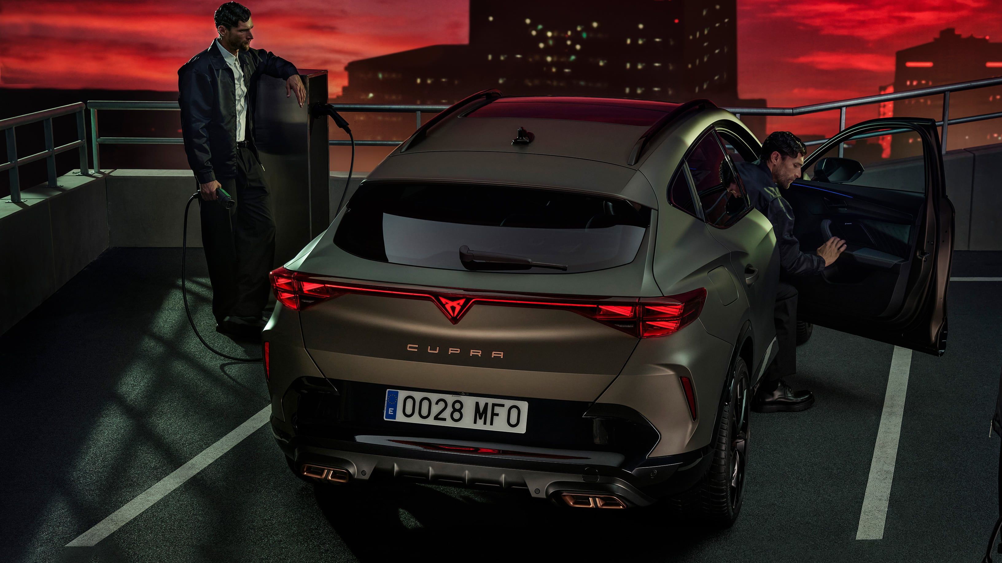 Bronasti avtomobil CUPRA Formentor PA je parkiran v garaži, medtem ko dve osebi stopata do vozila. Ena oseba zapira vrata avtomobila.