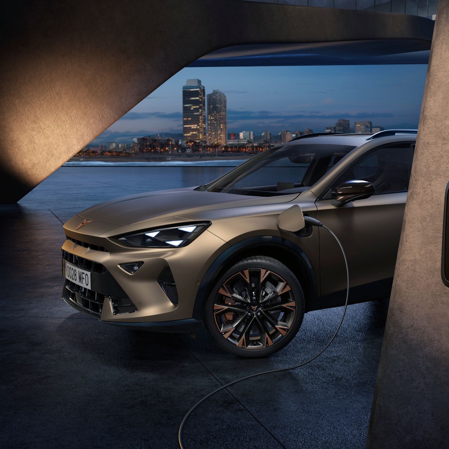 Električni CUPRA Formentor se polni na polnilni postaji ob sodobni stavbi. V ozadju je urbano mestno okolje z visokimi stavbami in obala. Avto je bronaste barve.